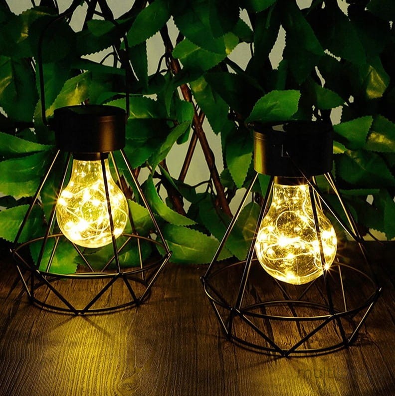 Czarna lampa stołowa wisząca Diament z metalową siatką, z widoczną żarówką LED emitującą ciepłe światło. Ujęcie z przodu, na drewnianym blacie, w otoczeniu zielonych liści. Lampy mają dekoracyjne, geometrzyczne kształty.