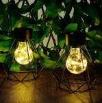 Czarna lampa stołowa wisząca Diament z metalową siatką, z widoczną żarówką LED emitującą ciepłe światło. Ujęcie z przodu, na drewnianym blacie, w otoczeniu zielonych liści. Lampy mają dekoracyjne, geometrzyczne kształty.