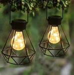 Czarna lampa stołowa wisząca Diament z metalową klatką, w której umieszczona jest żarówka z ozdobnymi światłami. Lampa zawieszona w otoczeniu zieleni, prezentująca nowoczesny design.