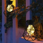 Lampa stołowa wisząca Diament w kolorze czarnym z przezroczystą żarówką LED w metalowej klatce, zawieszona w otoczeniu roślin. Oświetlenie emituje ciepłe światło, idealne do dekoracji przestrzeni na zewnątrz.
