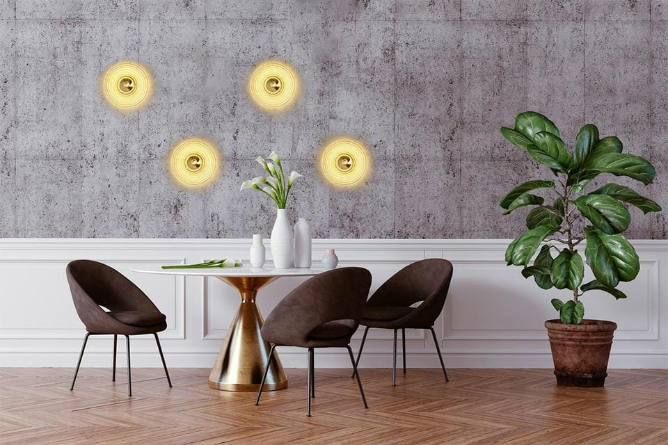 Kinkiet Onda LED 1xLED/9W w kolorze złotym, zamontowany na szarej ścianie. W tle widoczne nowoczesne krzesła i stół w aranżacji wnętrza, z rośliną doniczkową obok. Oświetlenie tworzy ciepłą atmosferę.