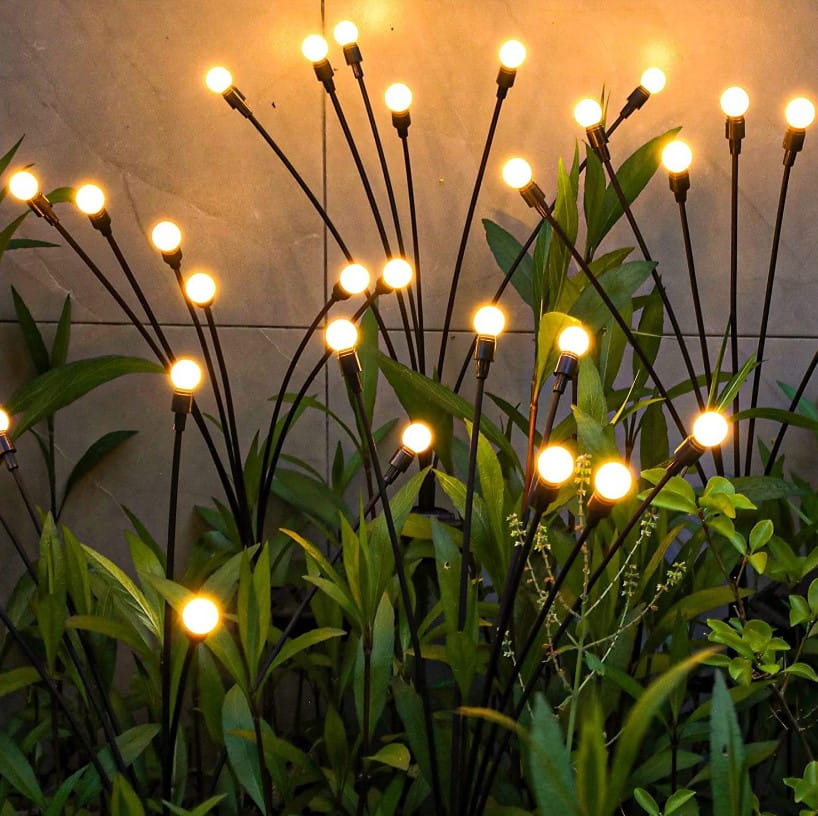 Lampa solarna ogrodowa 6xLED na czarnym szpikulcu, z sześcioma okrągłymi żarówkami emitującymi ciepłe światło, umieszczona wśród zielonych roślin. Ujęcie z boku, ukazujące delikatne światło w ogrodzie.