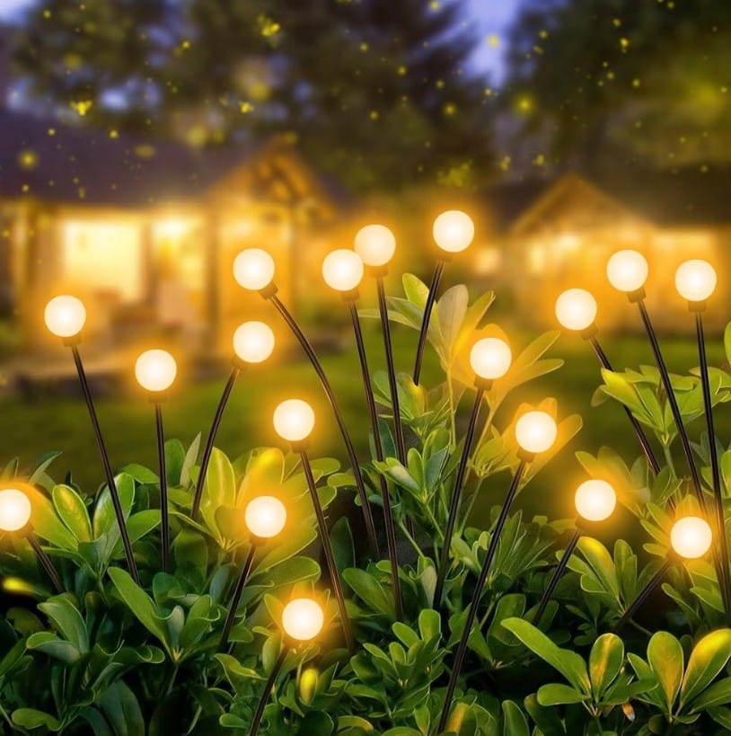 Lampa solarna ogrodowa z 6 diodami LED na czarnym szpikulcu, umieszczona w zielonym otoczeniu, świeci ciepłym światłem. Widoczna z góry na tle rozmytego, oświetlonego tła ogrodu.