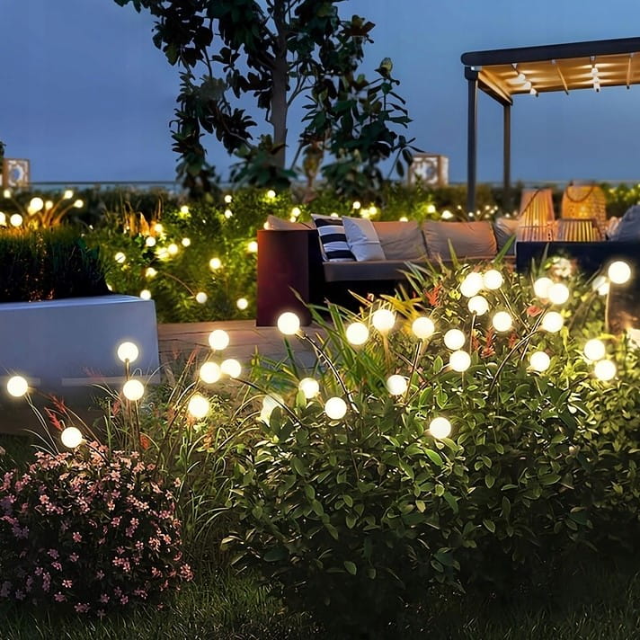 Lampa solarna ogrodowa 6xLED na szpikulcu w kolorze czarnym, umieszczona w zielonej roślinności. Widoczne delikatne, świecące kulki LED, które tworzą przyjemną atmosferę w ogrodzie. Tło z sofy i pergoli.