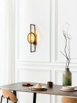 Kinkiet Troya w kolorze złotym z oprawą na jedną żarówkę E14, zamontowany na białej ścianie. Spherical klosz w eleganckiej ramie, oświetlający stół z ceramicznymi naczyniami i dekoracjami w aranżacji wnętrza.