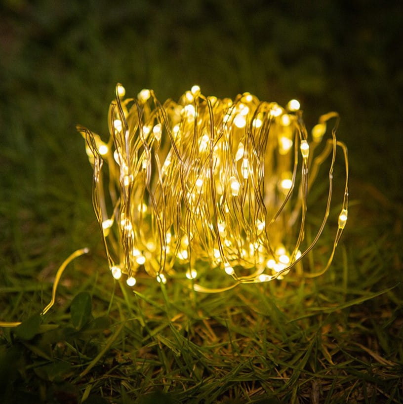 Girlanda świetlna ogrodowa solarna z 100 diodami LED, długość 10 m. Złote, cienkie przewody w kształcie spirali, umieszczone na zielonej trawie. Produkt leży na ziemi, w otoczeniu naturalnego krajobrazu, idealny do dekoracji ogrodu.