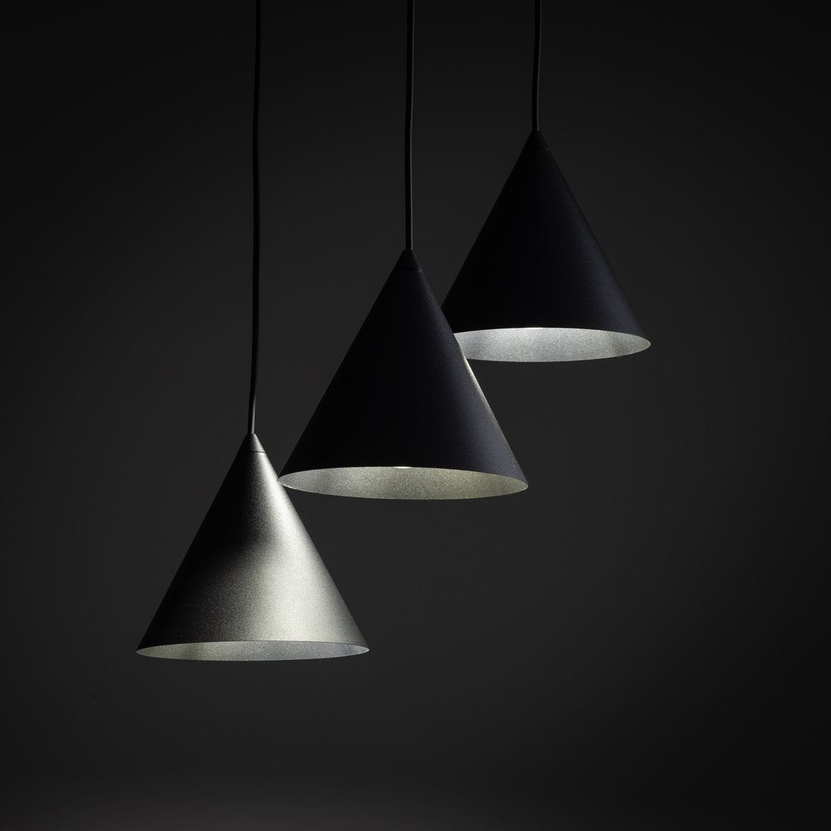 Lampa wisząca Cono z trzema kloszami w kształcie stożków, w kolorze czarnym i szarym, zawieszona na cienkich linkach. Klosze mają matową fakturę, a ich wnętrza są jasne, co podkreśla ich nowoczesny design.