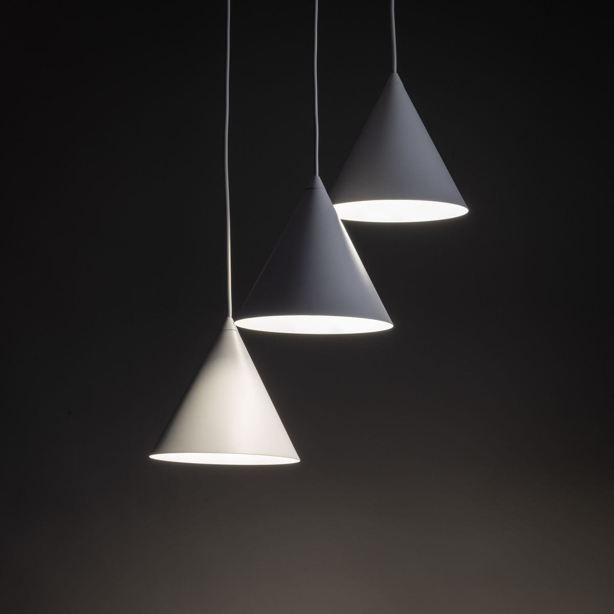 Lampa wisząca Cono z trzema stożkowymi kloszami w białym kolorze. Klosze zawieszone na białych przewodach, emitujące jasne światło. Ujęcie z przodu, na ciemnym tle.