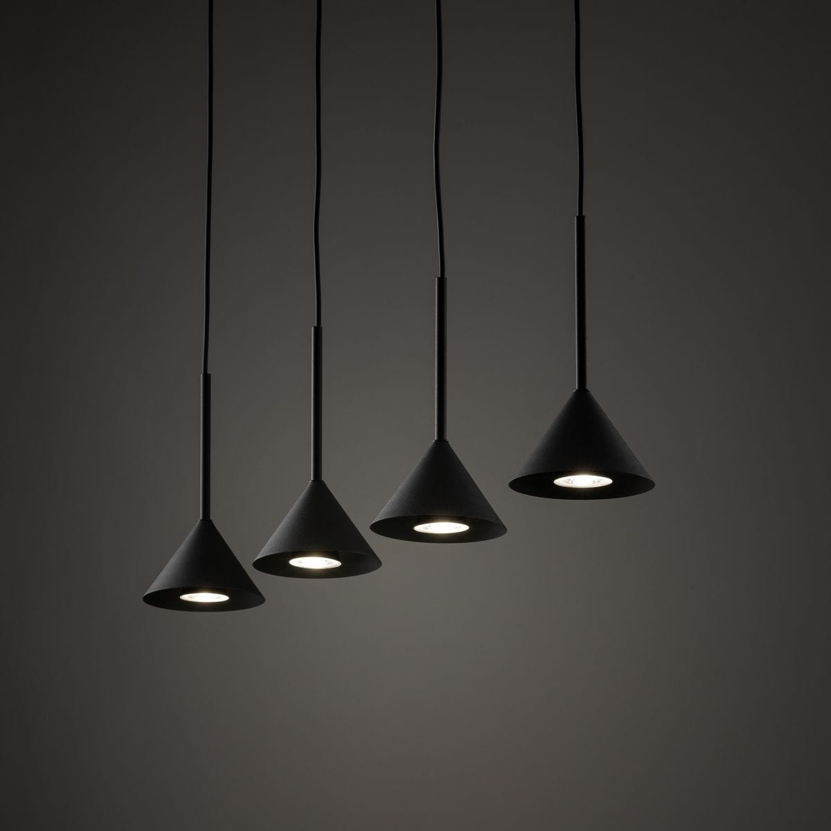 Czarna lampa wisząca Cono z czterema kloszami w kształcie stożka, zawieszona na cienkich kablach. Klosze mają matową powierzchnię i są ustawione w linię, emitując światło z żarówek GU10. Tło jest ciemne, co podkreśla elegancki wygląd lampy.