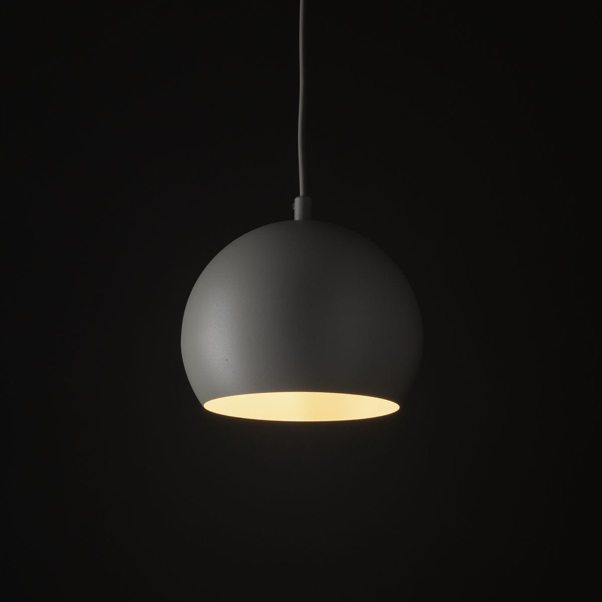 Lampa wisząca Zoe D17 w beżowym kolorze, o okrągłym kształcie, zawieszona na białym przewodzie. Emituje ciepłe światło przez otwór na dole. Ujęcie z przodu, na jednolitym, ciemnym tle.