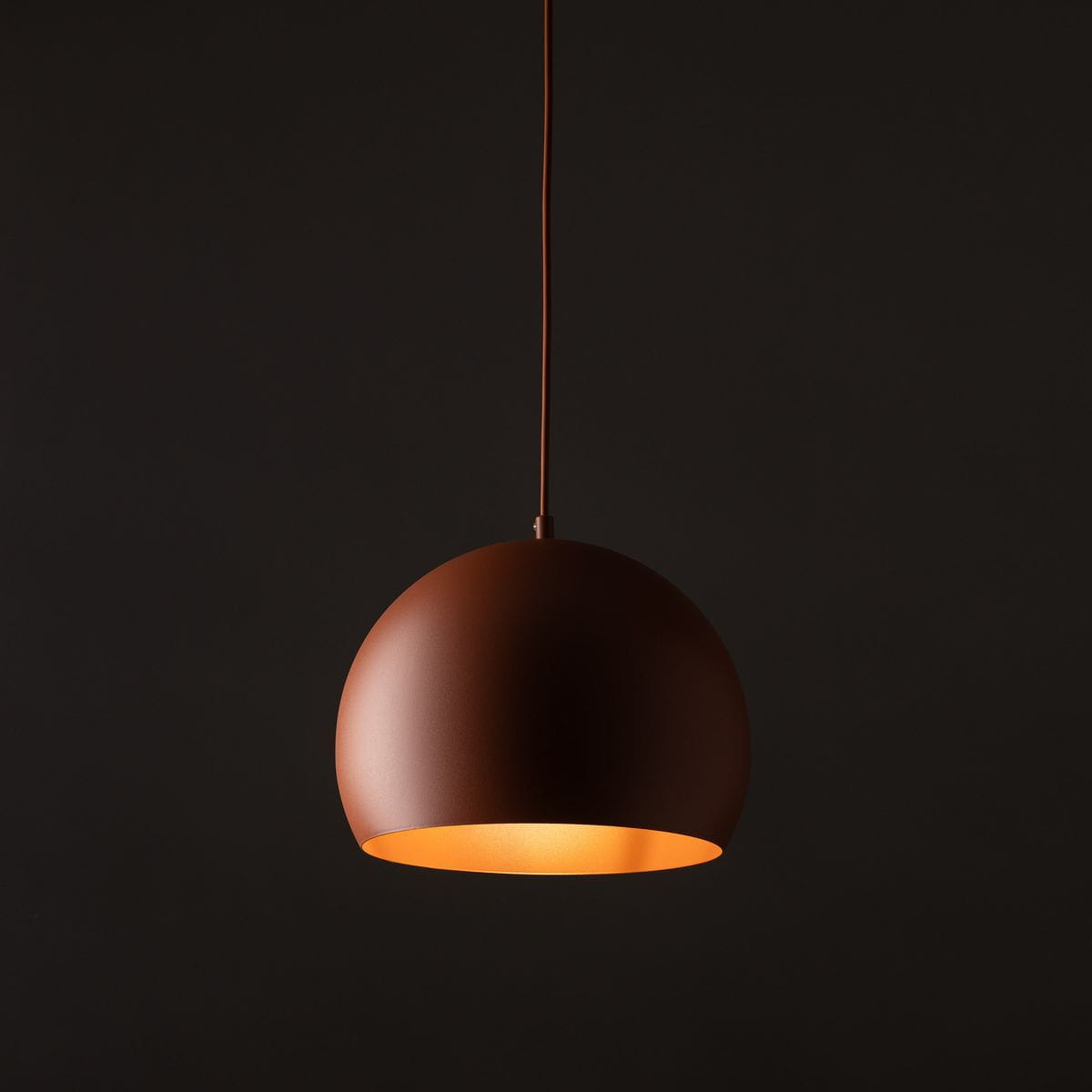 Lampa wisząca Zoe D27 w kolorze ceglastym z matowym wykończeniem, o kształcie półkuli. Zawieszona na cienkim przewodzie, emitująca ciepłe światło z wewnętrznej strony klosza. Idealna do nowoczesnych wnętrz.
