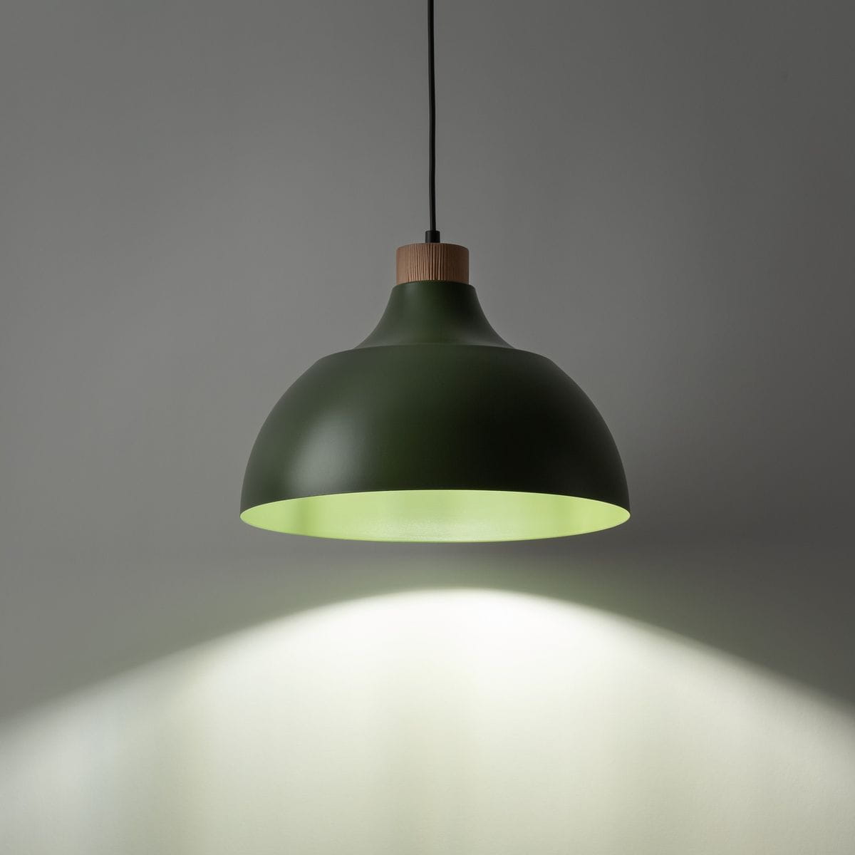 Lampa wisząca Cap w kolorze zielonym z metalowym kloszem o zaokrąglonym kształcie, z drewnianym elementem u góry. Oświetlenie skierowane w dół, stworzone dla żarówki E27. Tło w neutralnym odcieniu szarości.