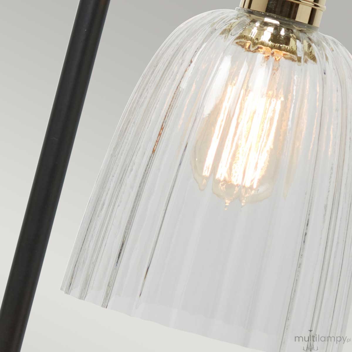 Lampka stołowa z czarną podstawą, posiada szklany klosz w formie cylindrycznej, z wyraźnymi żłobieniami. Widoczna żarówka E27 w środku. Ujęcie z boku, skupione na detalu klosza i źródle światła.