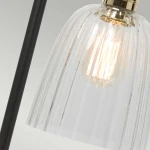 Lampka stołowa z czarną podstawą, posiada szklany klosz w formie cylindrycznej, z wyraźnymi żłobieniami. Widoczna żarówka E27 w środku. Ujęcie z boku, skupione na detalu klosza i źródle światła.