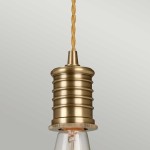 Lampa wisząca z postarzanym mosiądzu, z oprawą na żarówkę E27. Kształt cylindryczny z teksturą rowków, zawieszona na plecionym przewodzie w kolorze żółtym. Ujęcie zbliżeniowe, ukazujące detal i materiał.