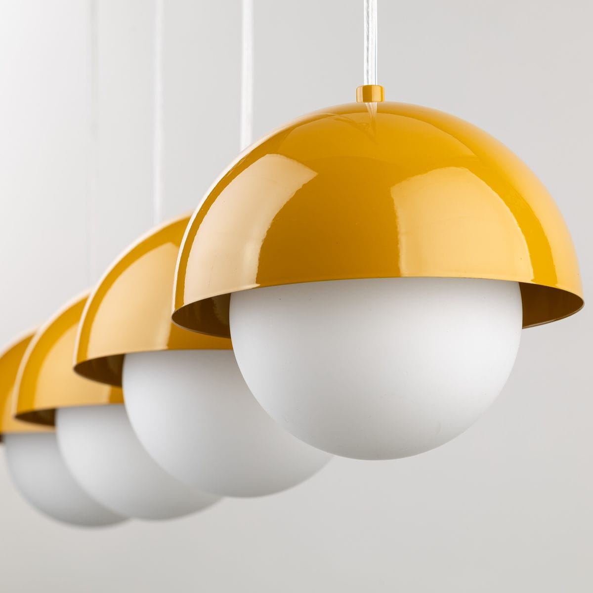 Lampa wisząca Bono 4xG9 w kolorze ochry z czterema zaokrąglonymi kloszami w białym kolorze, zawieszona na białych linkach. Klosze mają połyskujące żółte, metalowe osłony w formie półkul. Produkt widoczny z przodu, na jednolitym tle.