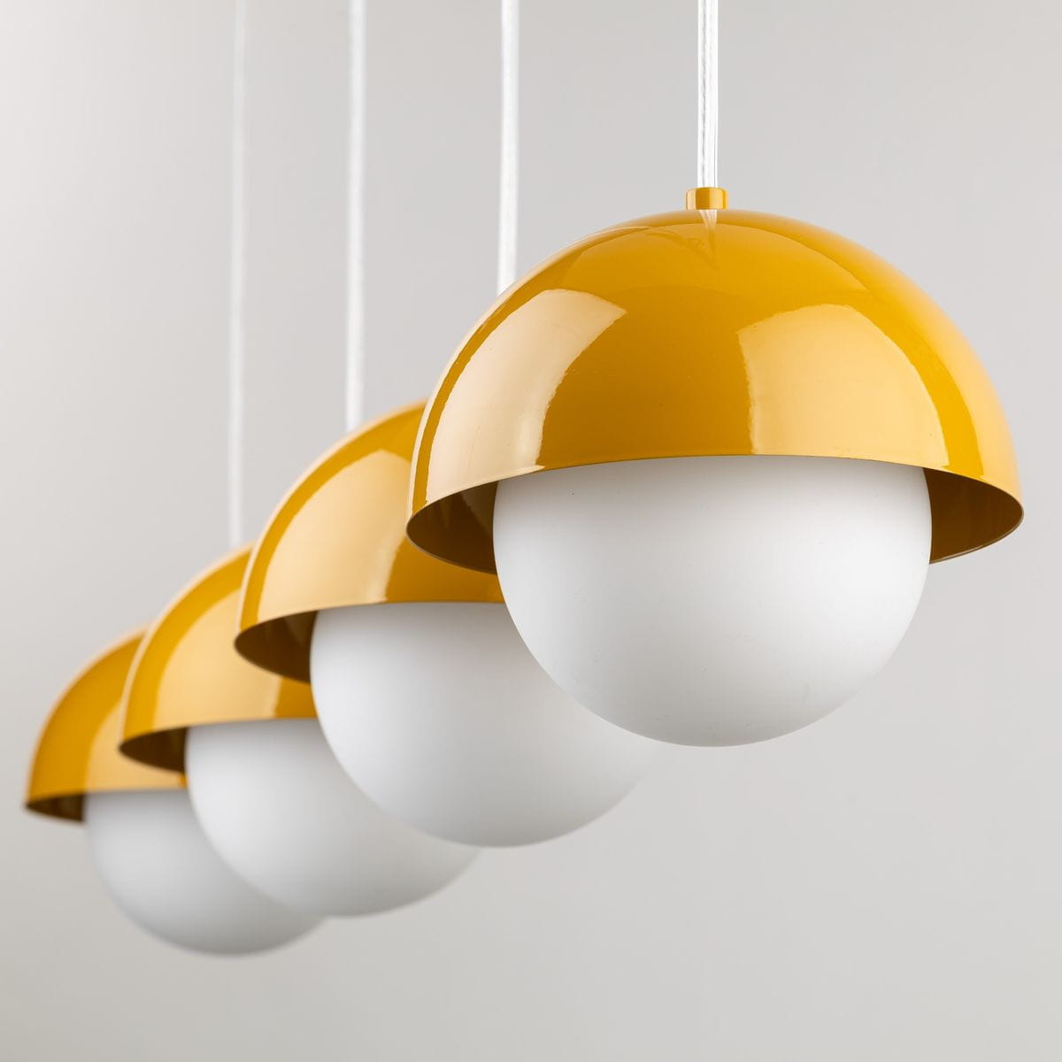 Lampa wisząca Bono z czterema kloszami w kolorze ochry, z białymi, okrągłymi kulami. Klosze mają połyskującą powierzchnię, zawieszone na białych linkach. Ujęcie z przodu, pokazujące równo rozmieszczone elementy.