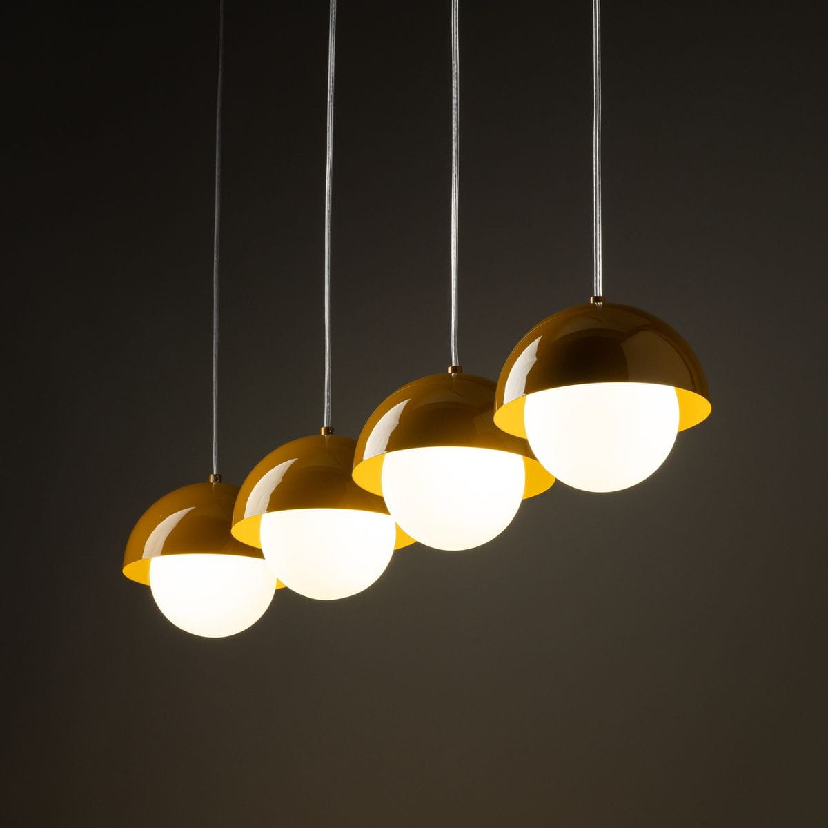 Lampa wisząca Bono z 4 kloszami w kolorze ochry, o półkulistym kształcie, zawieszona na białych linkach. Klosze o białych, matowych wnętrzach, emitujące ciepłe światło, prezentują się na ciemnym tle.