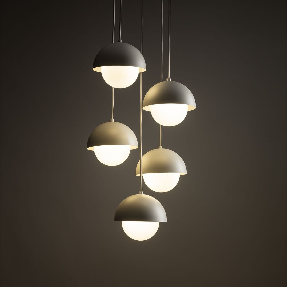 Beżowa lampa wisząca Bono z pięcioma kloszami w kształcie półkul, zawieszona na cienkich przewodach. Klosze mają białe, szklane wnętrza, emitujące miękkie światło. Lampa wisi w pionie, na ciemnym tle.