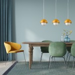 Lampa wisząca Bono D17 z jednym źródłem światła G9, w kolorze ochry, z zaokrąglonym kloszem połączonym z białą kulą. Zawieszona nad drewnianym stołem, w otoczeniu zielonych krzeseł i żółtego fotela. Tło w pastelowym odcieniu niebieskiego.