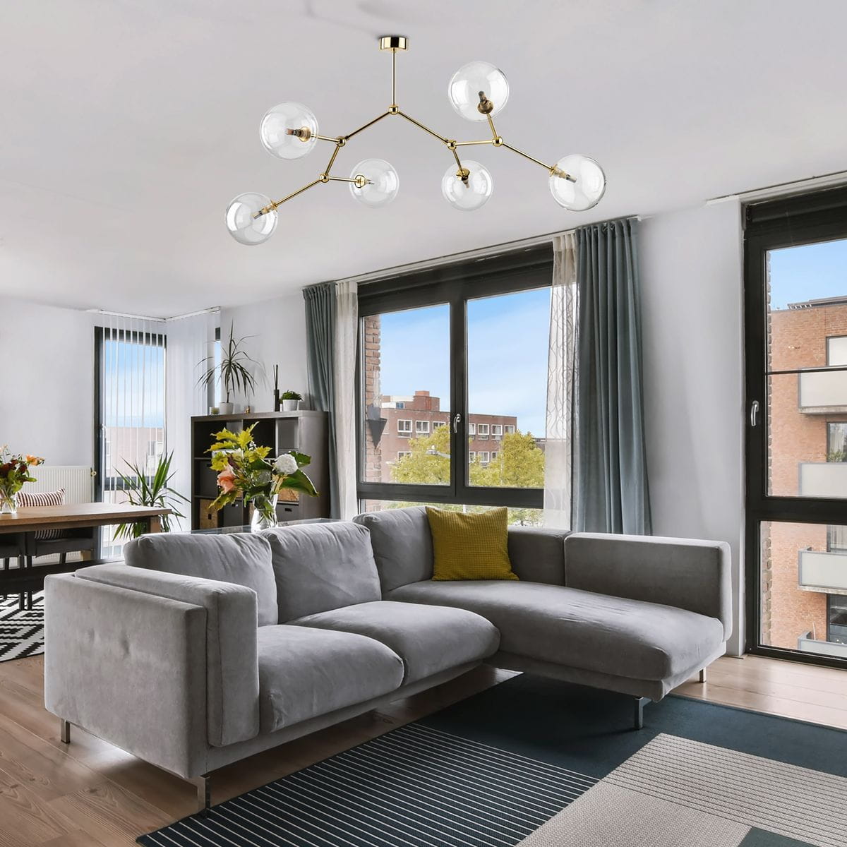 Lampa sufitowa Fairy w kolorze złotym i transparentnym, z sześcioma kulistymi kloszami na ramionach. Zamontowana na suficie, w nowoczesnym wnętrzu z szaro-białymi elementami. Widoczna w aranżacji z kanapą i oknami.