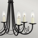 Lampa wisząca Minster w kolorze czarnym, z sześcioma ramionami na żarówki E14. Górna część z prostymi liniami, a dolne ramiona z dekoracyjnymi kręceniami. W lampie widoczne przezroczyste klosze w kształcie łzy oraz kremowe podstawy.