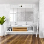 Kinkiet LED Isla IP44 w chromie, zamontowany nad lustrem w nowoczesnej łazience. Oświetlenie w formie prostokątnej listwy, emitujące białe światło, w otoczeniu białych umywalek i drewnianego blatu. Tło w odcieniach szarości z rośliną w donicy.