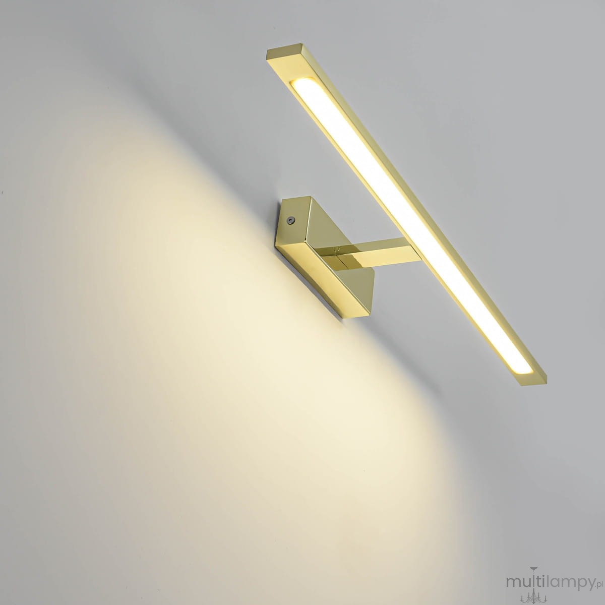 Kinkiet LED Isla IP44 w kolorze złotym, o wymiarach L62 cm i mocy 20W. Prosty, prostokątny kształt z zamontowanym źródłem światła, emitującym ciepłe światło. Montowany na ścianie, w tle jasna powierzchnia.