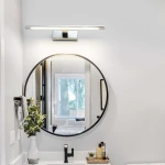 Kinkiet LED Isla IP44 w kolorze chromu, zamontowany nad okrągłym lustrem. Posiada prostą, smukłą formę oraz świeci ciepłym światłem. Widoczne detale to chromowane wykończenie i minimalistyczny design, idealny do nowoczesnych wnętrz.