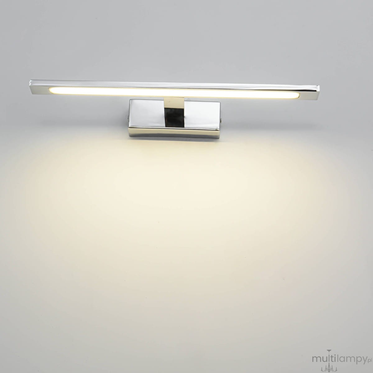 Kinkiet LED Isla IP44 w kolorze chromu, o prostokątnym kształcie, zamontowany na ścianie. Lampa emituje ciepłe światło, widoczna podstawa i wąska, płaska oprawa świetlna. Tło jasne, jednolite.