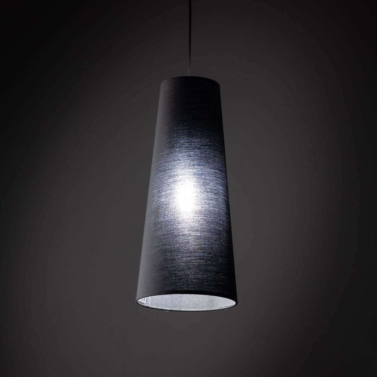 Lampa wisząca Zing w kształcie stożka, czarna, z materiału tekstylnego. Ujęcie z dołu, z widoczną oprawką na żarówkę E27. Wewnątrz lampa emituje jasne światło, tło ciemne, podkreślające elegancki design.