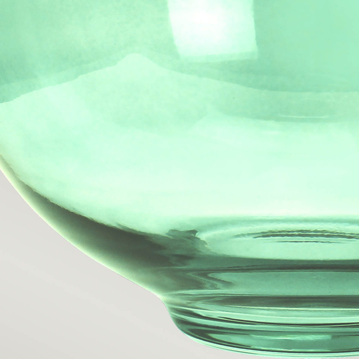 Zielona lampka stołowa Kara z kloszem w odcieniu pastelowej zieleni, wykonana ze szkła. Ujęcie zbliżeniowe na dolną część klosza, ukazujące gładką fakturę i zaokrąglony kształt. Tło jednolite, jasne, co podkreśla kolor lampki.