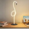 Lampka stołowa w kształcie flaminga, wykonana z białego materiału, z LED 6W. Posiada czarną podstawę i wygięty kształt przypominający szyję ptaka. Ujęcie frontalne na tle jasnej ściany, z książkami obok.