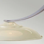 Lampka stołowa Franklin w kolorze kremowym z metalowym, lekko zakrzywionym uchwytem. Widoczna ceramiczna podstawa oraz oprawa na żarówkę E27. Ujęcie z boku, ukazujące detale kształtu i materiału. Tło jednolite, szare.
