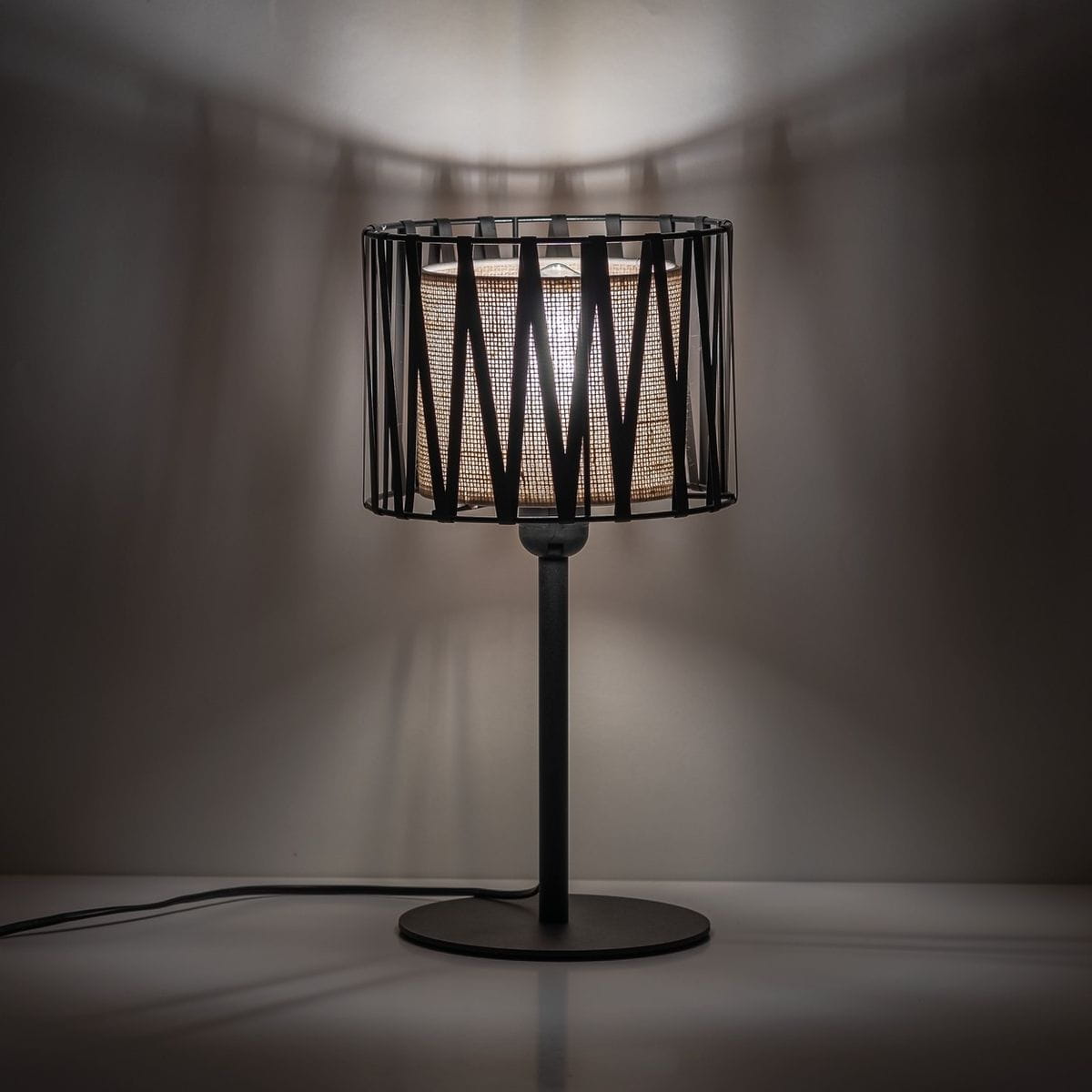 Lampka stołowa Harmony w kolorze czarnym i brązowym z metalowym kloszem w geometrycznym wzorze. Stoi na okrągłej podstawie, z widoczną żarówką E27. Tło jednolite, podkreślające design lampy.