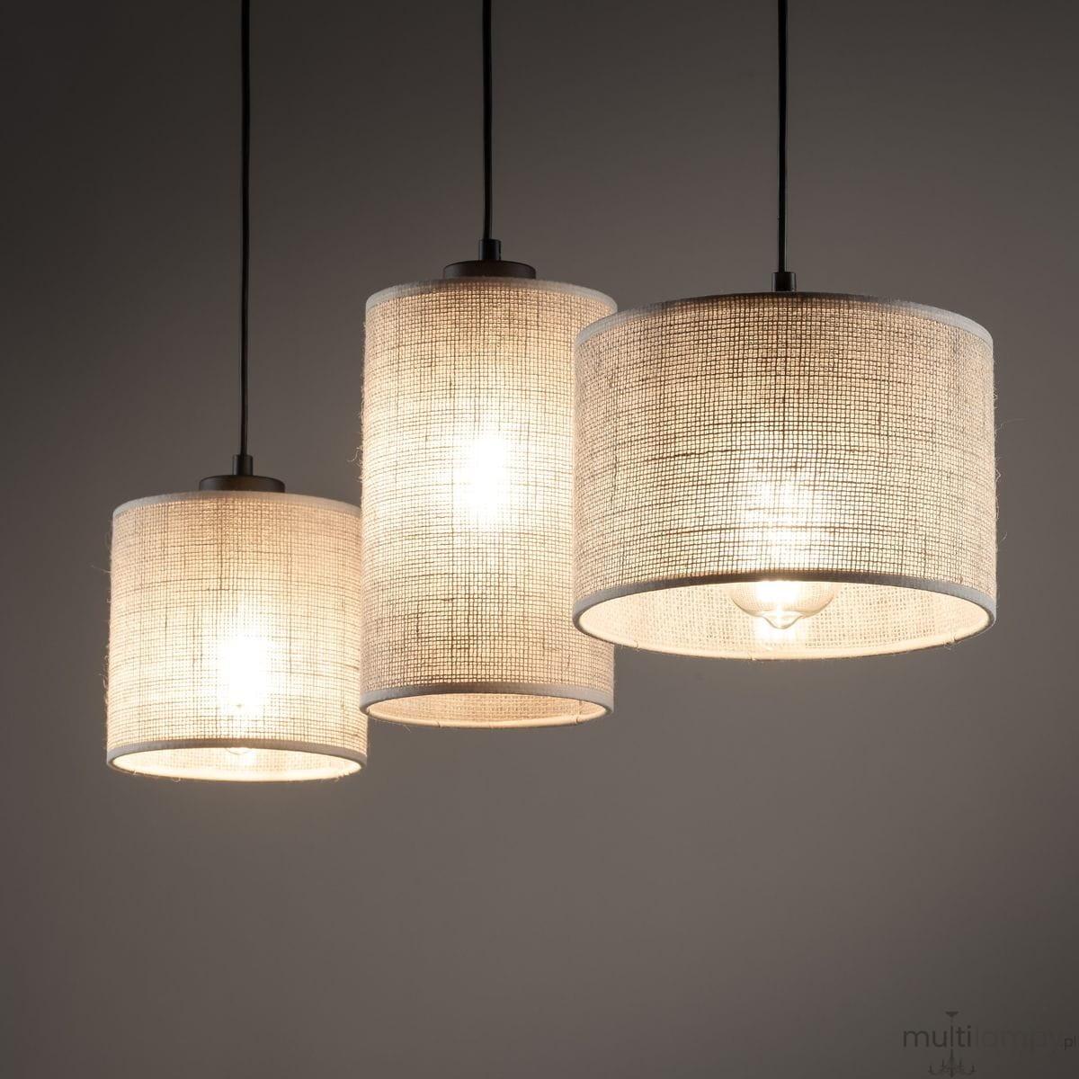 Lampa wisząca Juta z trzema abażurami w kolorze naturalnym z juty, zawieszona na czarnych kablach. Abażury mają okrągły kształt, a każdy z nich jest podświetlony, tworząc ciepłe światło. Tło ciemnoszare, minimalistyczne.