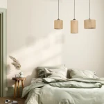 Lampa wisząca Juta z trzema kloszami w kolorze czarnym i brązowym, zawieszona nad łóżkiem, w otoczeniu pastelowych poduszek i narzuty. Klosze wykonane z materiału, w kształcie cylindrów, na czarnych przewodach.