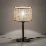 Lampka stołowa z okrągłym kloszem z naturalnej juty w odcieniu brązowym, na czarnej metalowej podstawie. Ustawiona na stole, z przewodem elektrycznym widocznym z boku. Klosz emitujący ciepłe światło, nadający przytulny klimat.