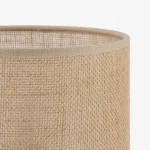 Lampka stołowa z juty w kolorze brązowym, z jasnym obrzeżem. Widoczny teksturowany klosz o cylindrycznym kształcie. Produkt umieszczony na jednolitym tle, bez widocznych źródeł światła.
