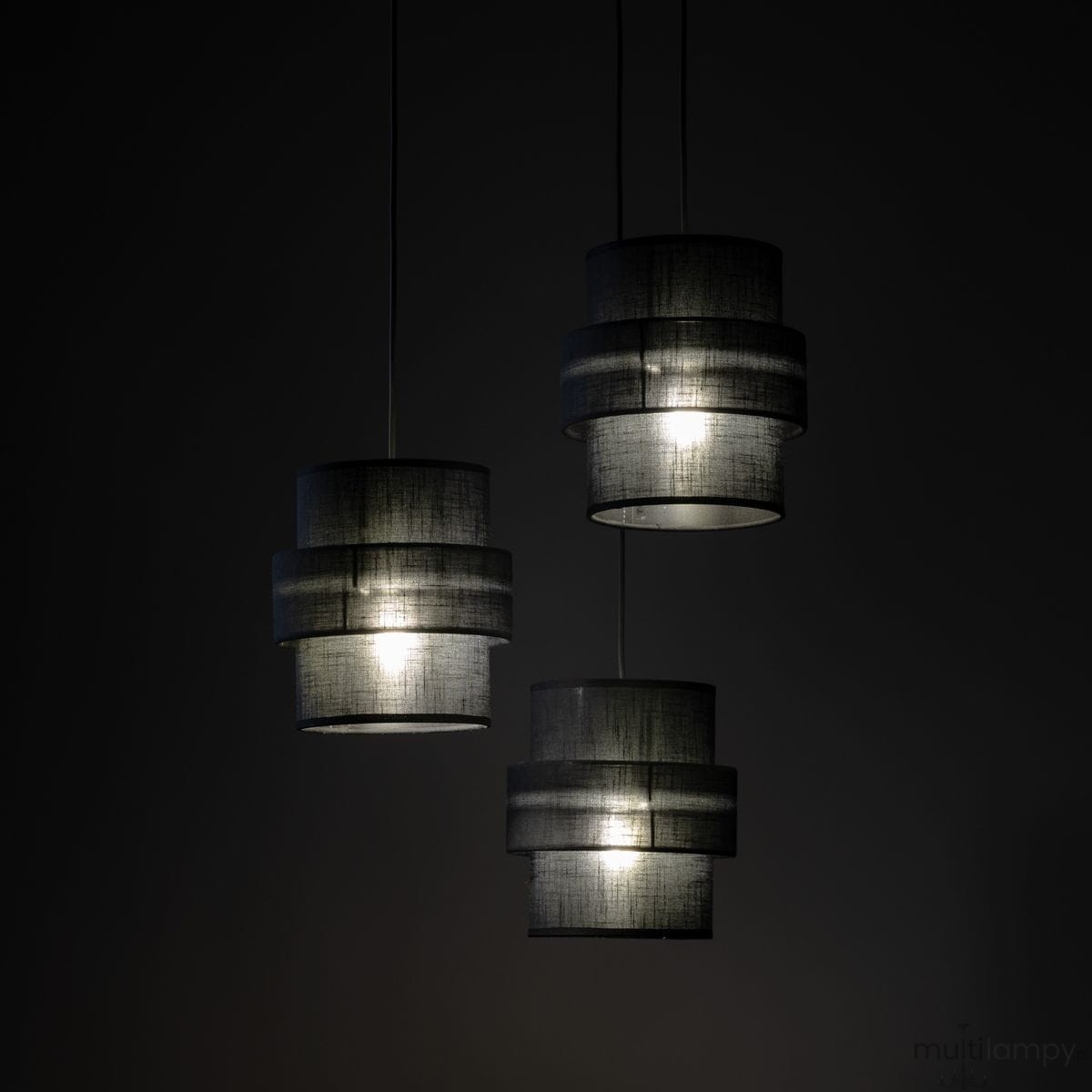 Lampa wisząca Calisto w kolorze czarnym z trzema abażurami, każdy w formie cylindrycznej, umieszczona na długich linkach. Wnętrza abażurów są jasne, co podkreśla światło. Produkt zawiera miejsce na trzy żarówki E27. Tło ciemne.