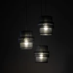 Lampa wisząca Calisto w kolorze czarnym z trzema abażurami, każdy w formie cylindrycznej, umieszczona na długich linkach. Wnętrza abażurów są jasne, co podkreśla światło. Produkt zawiera miejsce na trzy żarówki E27. Tło ciemne.