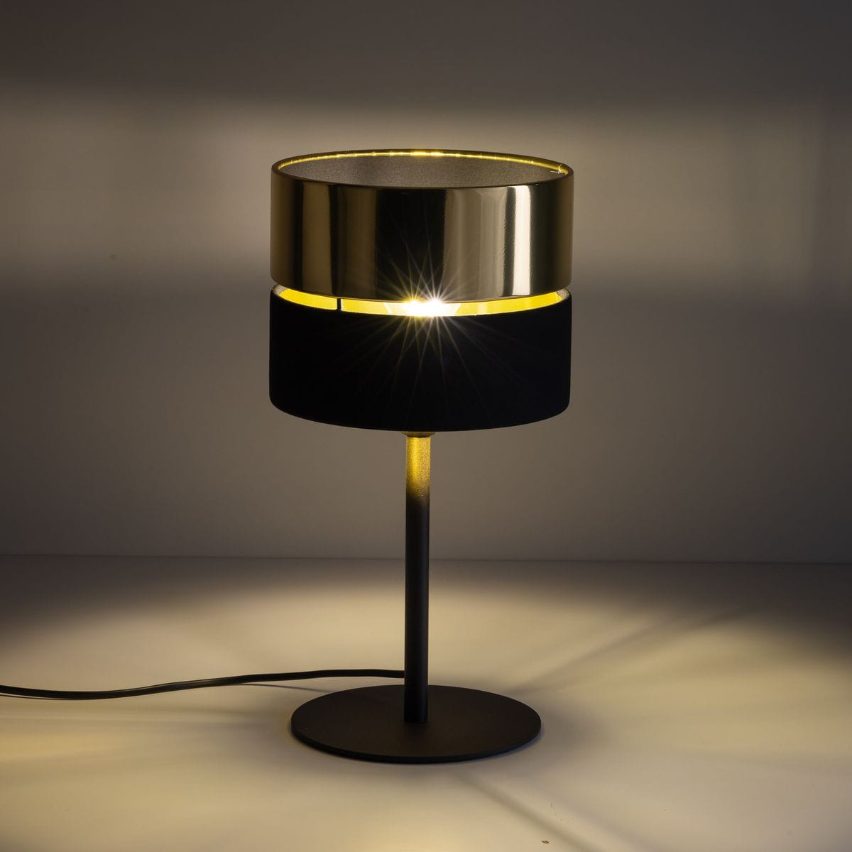 Lampa stołowa Hilton 1xE27 w kolorze czarnym i złotym, z okrągłym kloszem, umieszczona na smukłej, metalowej nodze. Klosz ma wykończenie połyskliwe, a dolna część z czarnego materiału.