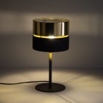Lampa stołowa Hilton 1xE27 w kolorze czarnym i złotym, z okrągłym kloszem, umieszczona na smukłej, metalowej nodze. Klosz ma wykończenie połyskliwe, a dolna część z czarnego materiału.
