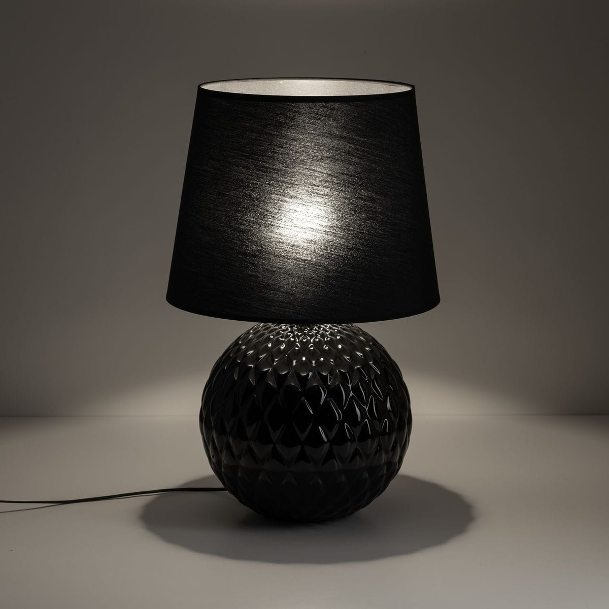 Lampa stołowa Santana z czarnym kloszem i dekoracyjną, teksturowaną podstawą w kształcie kuli. Ujęcie frontalne, lampa stoi na białym tle, podkreślając jej elegancki design. Przewód zasilający widoczny z boku.