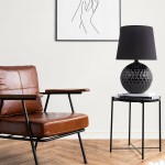 Lampa stołowa Santana 1xE27 w kolorze czarnym, z okrągłą, teksturowaną podstawą i prostym, czarnym kloszem. Ustawiona na czarnym stoliku obok brązowego fotela. Tło w jasnym kolorze, z obrazem na ścianie.