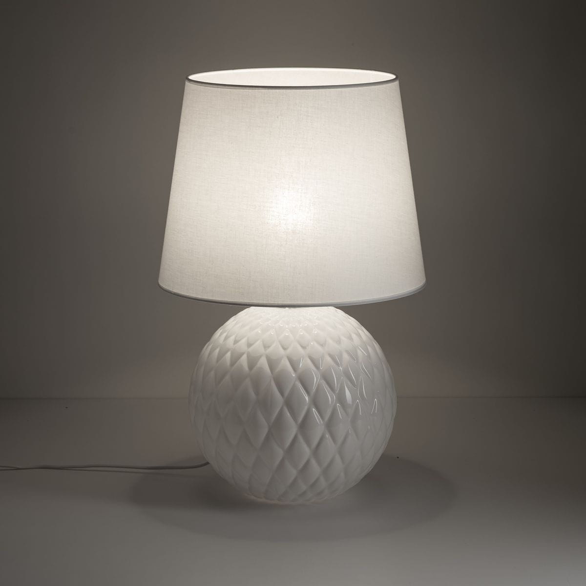 Lampa stołowa Santana z białym, tekstylnym kloszem i kulistą podstawą z fakturowanej ceramiki. Ujęcie z boku, ukazujące detale wzoru na podstawie oraz delikatne światło wydobywające się z klosza.