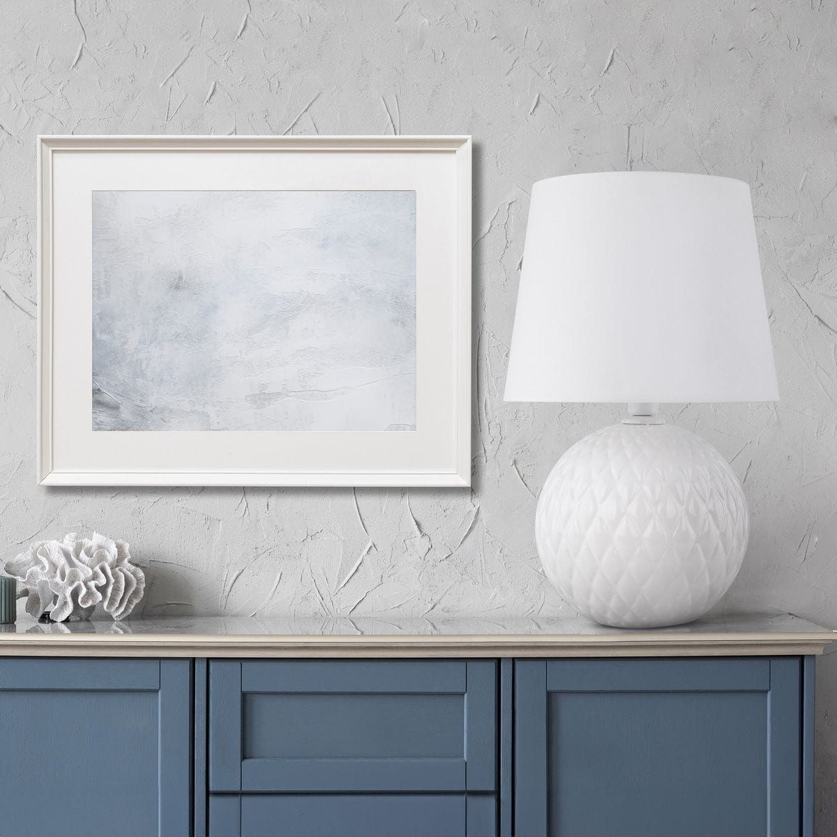 Lampa stołowa Santana z białym, okrągłym kloszem i fakturowanym, ceramicznym stelażem w kształcie kuli. Ustawiona na niebieskiej komodzie, obok dekoracji. Klosz w prostym, eleganckim stylu, idealna do nowoczesnych wnętrz.