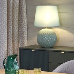 Lampa stołowa Santana w kolorze miętowym z ceramiczną, żebrowaną podstawą i materiałowym kloszem. Ujęcie frontalne, lampa stoi na ciemnozielonej szafce obok szklanek i dzbanka. Tło z jasnymi zasłonami i drewnianą ścianą.