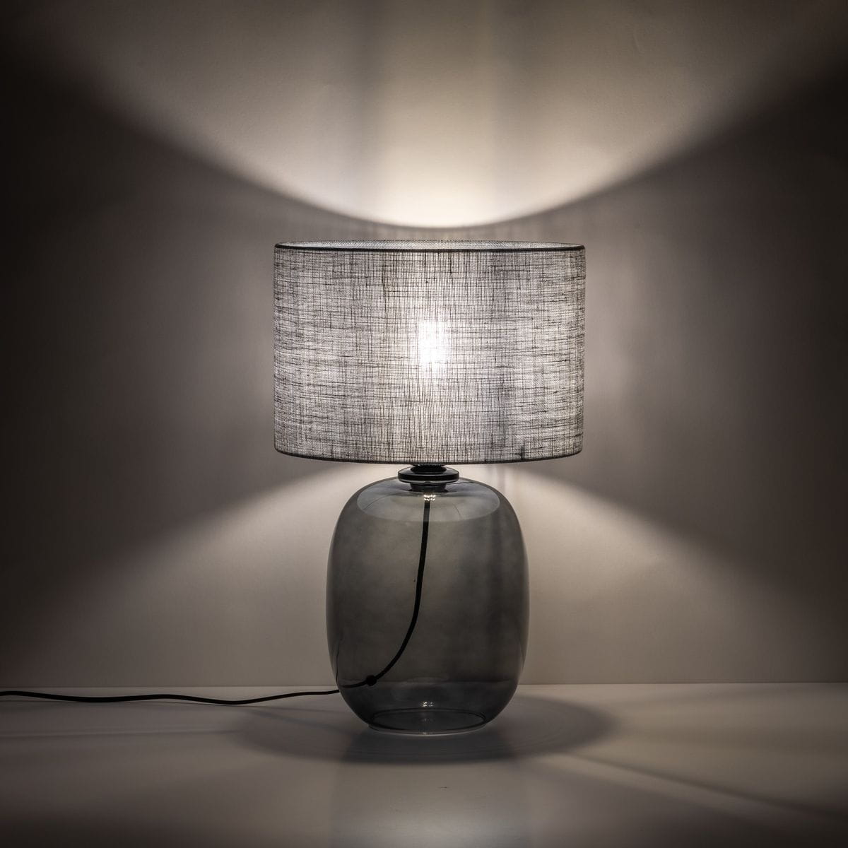 Lampa stołowa Melody z grafitowym, szklanym korpusem i szarym, tekstylnym kloszem. Ujęcie z boku, eksponujące okrągły kształt podstawy oraz detale materiału klosza. Widoczny kabel zasilający, delikatne światło tworzy cienie na tle.