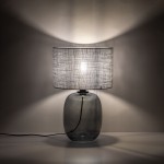 Lampa stołowa Melody z grafitowym, szklanym korpusem i szarym, tekstylnym kloszem. Ujęcie z boku, eksponujące okrągły kształt podstawy oraz detale materiału klosza. Widoczny kabel zasilający, delikatne światło tworzy cienie na tle.